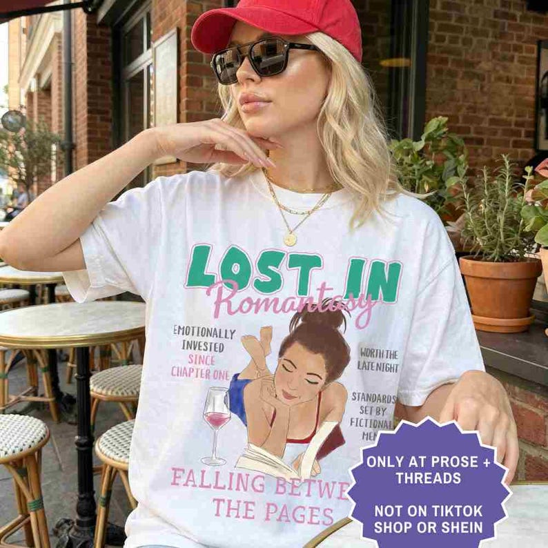 May include: White t-shirt with the text "LOST IN Romantsiy" in green and pink. The shirt features an illustration of a woman reading a book with a glass of wine. Additional text includes "Emotionally Invested Since Chapter One" and "Falling Between The Pages."
