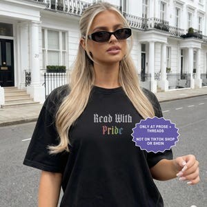 Pu&ograve; includere: Una t-shirt nera a maniche corte con il testo Read With Pride in un carattere gotico, con la parola Pride visualizzata nei colori dell'arcobaleno. Un grafico viola a forma di stella include il testo ONLY AT PROSE + THREADS NOT ON TIKTOK SHOP OR SHEIN.