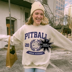 May include: A beige sweatshirt with a blue and white graphic print that says "Pitball" and "Zodiac Academy". The graphic also includes a wreath with the words "Solarbia Champs" and the number "20". A black star with the text "Prose + Threads" is in the upper right corner of the image.