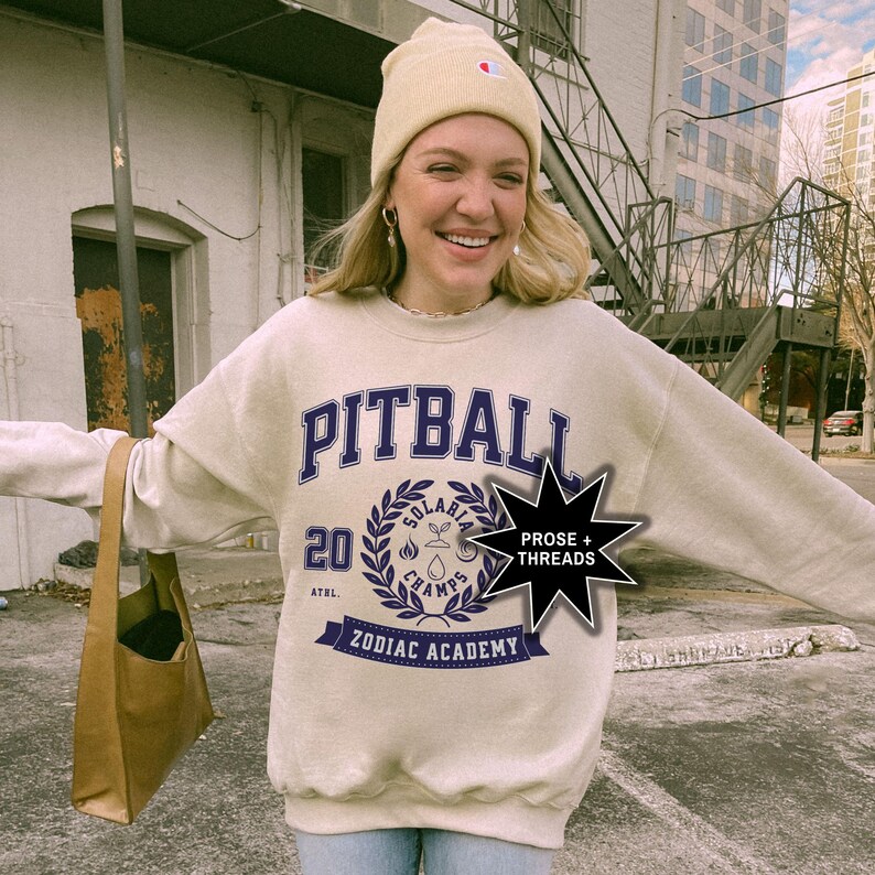 May include: A beige sweatshirt with a blue and white graphic print that says "Pitball" and "Zodiac Academy". The graphic also includes a wreath with the words "Solarbia Champs" and the number "20". A black star with the text "Prose + Threads" is in the upper right corner of the image.