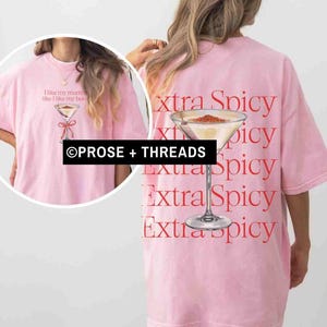 May include: A light pink t-shirt with a martini glass graphic and the text "I like my martinis like I like my boobs" on the front. The back of the shirt features the words "extra spicy" and a martini glass.