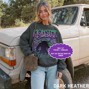 May include: Dark heather gray sweatshirt with the text "Monster Romance Book Club" in green and purple. The graphic features a silhouette of a horned creature. A small, patterned purse hangs from a chain.