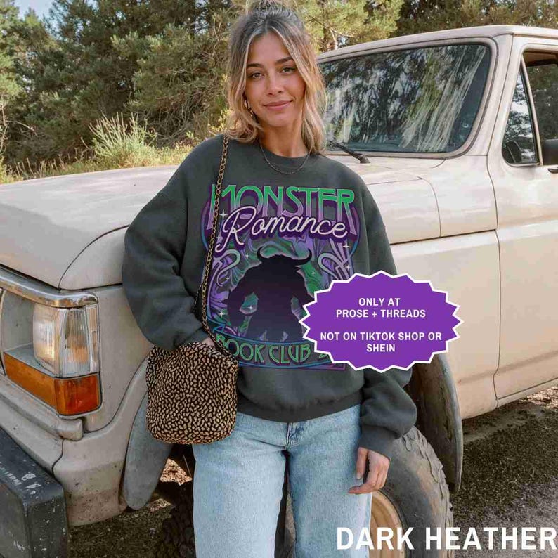 May include: Dark heather gray sweatshirt with the text "Monster Romance Book Club" in green and purple. The graphic features a silhouette of a horned creature. A small, patterned purse hangs from a chain.
