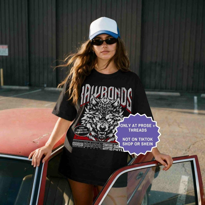 Op de afbeelding: Zwart T-shirt met een wolfafbeelding en de tekst "IAWBONDS". Het shirt heeft ook de tekst "LIVE FROM THE BONDED CITY" en "ADVISORY". Het shirt wordt gedragen met een wit en blauw baseballpetje en een zonnebril.