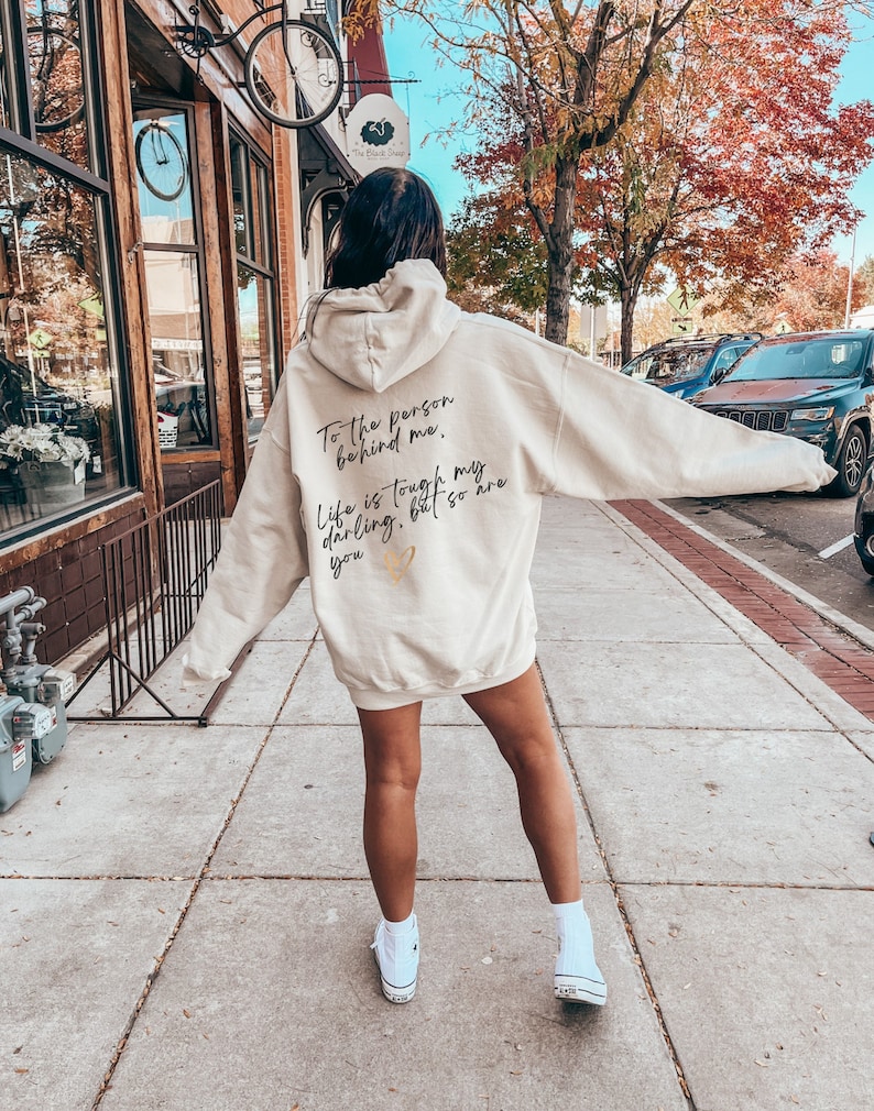May include: Cream-colored hoodie with the text "To the person behind me. Life is tough my darling, but so are you" in black script. A small gold heart is below the text. The hoodie is worn with white high-top sneakers.