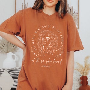 May include: Rust-colored t-shirt featuring a white graphic of Medusa with the text "HER WALLS WERE BUILT BY THE BETRAYALS of those she loved - MEDUSA-". The design is centered on the front of the shirt.