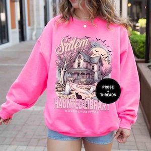 May include: A pink sweatshirt with a graphic print of a house with a spooky Halloween theme. The text on the sweatshirt says "Salem Haunted Library Massachusetts" and "Prose + Threads".
