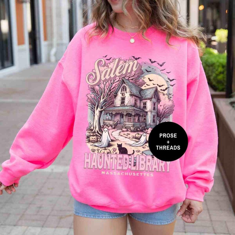 May include: A pink sweatshirt with a graphic print of a house with a spooky Halloween theme. The text on the sweatshirt says "Salem Haunted Library Massachusetts" and "Prose + Threads".