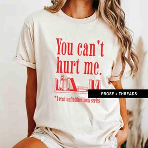 May include: A beige t-shirt with the text "You can't hurt me." in red. Below the text is a graphic of a bookshelf with books. The text "*I read unfinished book series" is below the graphic.