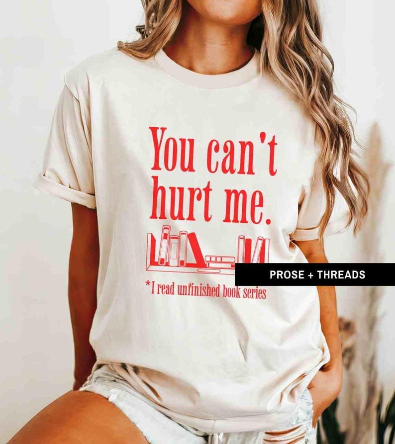May include: A beige t-shirt with the text "You can't hurt me." in red. Below the text is a graphic of a bookshelf with books. The text "*I read unfinished book series" is below the graphic.