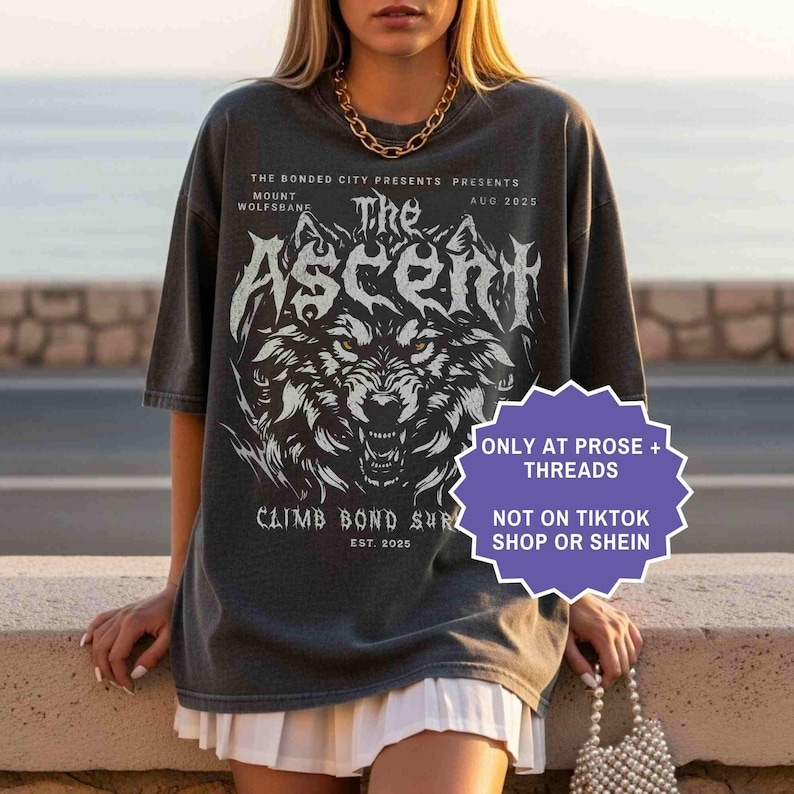 May include: Dark gray t-shirt featuring a wolf graphic with the text "The Ascent." Additional text includes "The Bonded City Presents," "Mount Wolfsbane," and "Climb Bond Sur." The shirt also has the text "Aug 2025" and "Est. 2025."