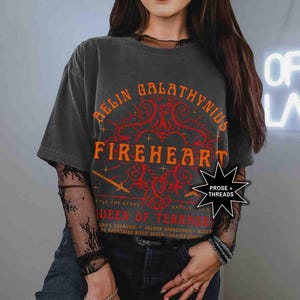 Pode incluir: Camiseta cinza escuro com as palavras "AELIN GALATHYNIUS FIREHEART" em laranja. A camiseta tamb&eacute;m tem o texto "QUEEN OF TERRASEN" e "PROSE + THREADS". A camiseta &eacute; usada com uma camisa de manga comprida de renda preta.