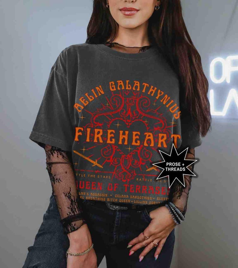 Pode incluir: Camiseta cinza escuro com as palavras "AELIN GALATHYNIUS FIREHEART" em laranja. A camiseta tamb&eacute;m tem o texto "QUEEN OF TERRASEN" e "PROSE + THREADS". A camiseta &eacute; usada com uma camisa de manga comprida de renda preta.