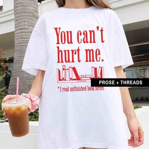 May include: White t-shirt with red text that says "You can't hurt me."  The text is above a graphic of a bookshelf with books on it. The text below the graphic says "*I read unfinished book series."