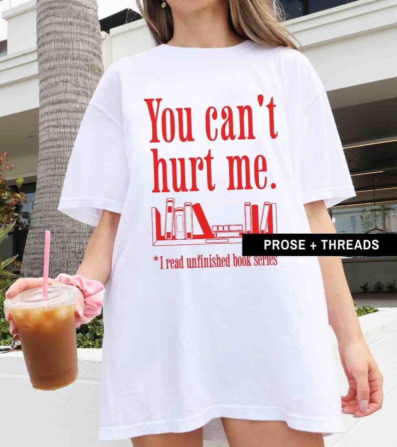 May include: White t-shirt with red text that says "You can't hurt me."  The text is above a graphic of a bookshelf with books on it. The text below the graphic says "*I read unfinished book series."