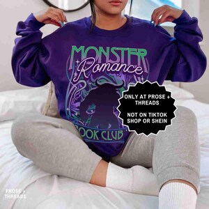 May include: A purple crewneck sweatshirt with the words "MONSTER Romance BOOK CLUB" in a stylized design. The graphic includes a silhouette of a horned figure. The sweatshirt has long sleeves.