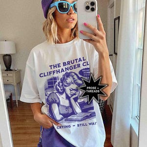 May include: White t-shirt with a purple and blue graphic that reads "THE BRUTAL CLIFFHANGER CLUB" and features a crying woman reading a book. The shirt is paired with purple shorts and a purple beanie. The person is wearing blue sunglasses.
