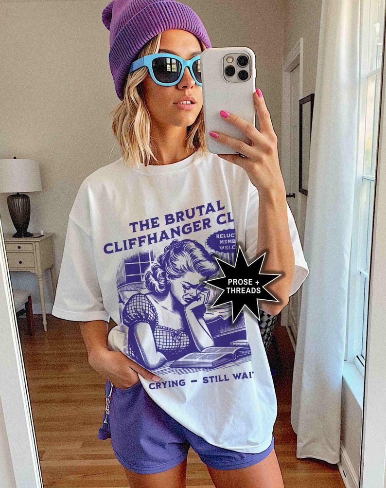 May include: White t-shirt with a purple and blue graphic that reads "THE BRUTAL CLIFFHANGER CLUB" and features a crying woman reading a book. The shirt is paired with purple shorts and a purple beanie. The person is wearing blue sunglasses.