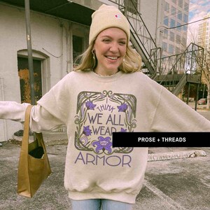 May include: A beige sweatshirt with a floral design and the text "I think we all wear armor". The sweatshirt has a crew neck and long sleeves. The model is wearing a beige beanie and blue jeans.
