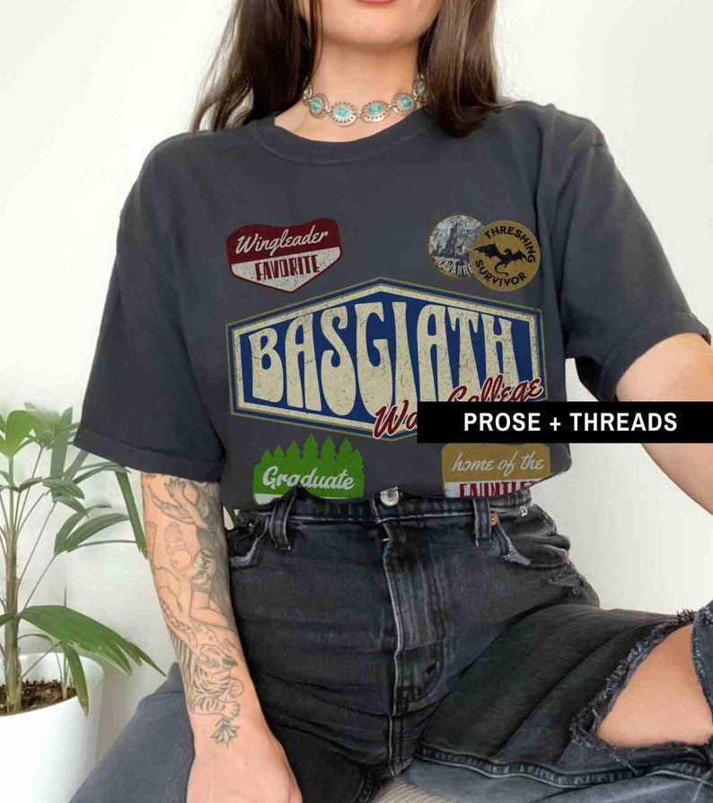 May include: A black t-shirt with a vintage-style graphic design featuring the text "Basgath" and various patches. The patches include "Wingleader Favorite", "Thresning Survivor", "Graduate", and "Home of the ...".