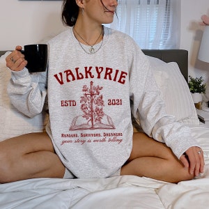 Może przedstawiać: Szara bluza z czerwonym nadrukiem przedstawiającym miecz, r&oacute;że i książkę. Tekst głosi "Valkyrie Est. 2021" i "Readers, Survivors, Dreamers your story is worth telling".