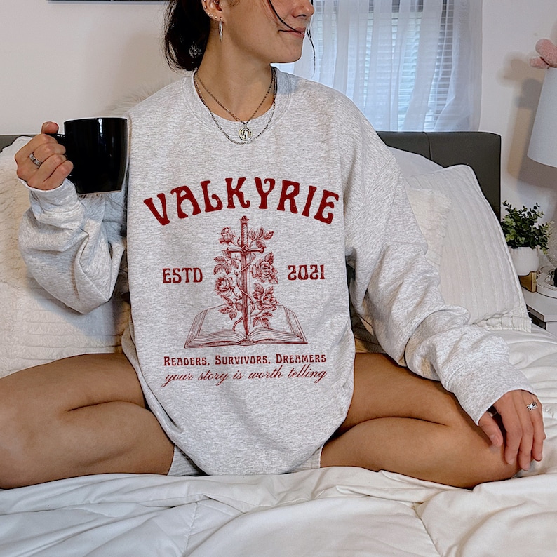 Może przedstawiać: Szara bluza z czerwonym nadrukiem przedstawiającym miecz, r&oacute;że i książkę. Tekst głosi "Valkyrie Est. 2021" i "Readers, Survivors, Dreamers your story is worth telling".