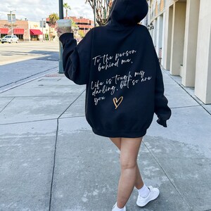May include: Black hooded sweatshirt with white and gold text on the back. The text reads "To the person behind me, Life is tough my darling, but so are you" with a gold heart. The sweatshirt is worn with white sneakers.