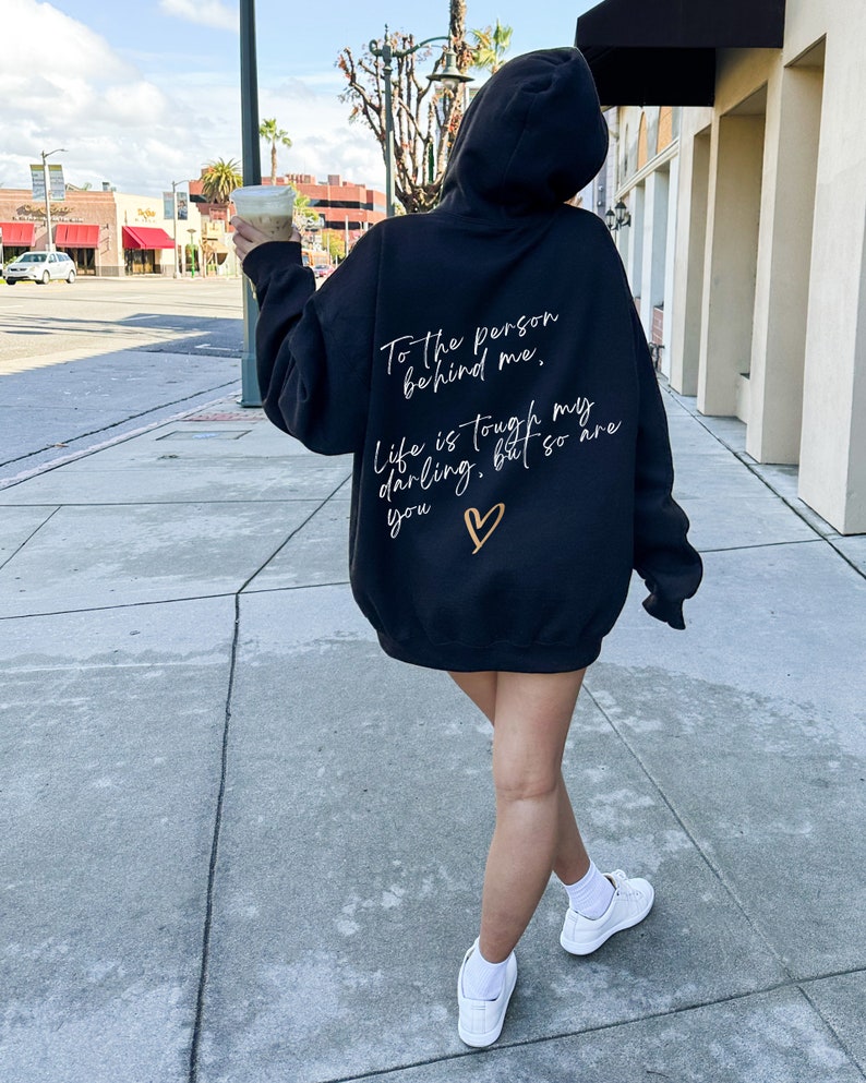 May include: Black hooded sweatshirt with white and gold text on the back. The text reads "To the person behind me, Life is tough my darling, but so are you" with a gold heart. The sweatshirt is worn with white sneakers.