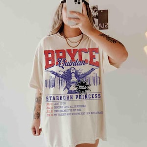 May include: A white t-shirt with a vintage-style graphic print featuring the text "Bryce Quinlan" in red and blue lettering. The graphic also includes a woman with outstretched arms, a cityscape, and the text "Starborn Princess". Below the graphic, there is a list of dates and phrases, including "Light it Up!" and "My Friends Are With Me And I Am Not Afraid."