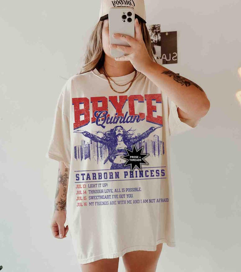 May include: A white t-shirt with a vintage-style graphic print featuring the text "Bryce Quinlan" in red and blue lettering. The graphic also includes a woman with outstretched arms, a cityscape, and the text "Starborn Princess". Below the graphic, there is a list of dates and phrases, including "Light it Up!" and "My Friends Are With Me And I Am Not Afraid."