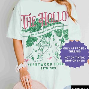 Pu&ograve; includere: T-shirt bianco con grafica verde e rosa con la scritta "The Hollow Inn for Travelers and Adventurers" con un disegno di casa sull'albero. La maglietta ha anche la scritta "Merrywood Forest ESTD 2021". La modella indossa la taglia XL.