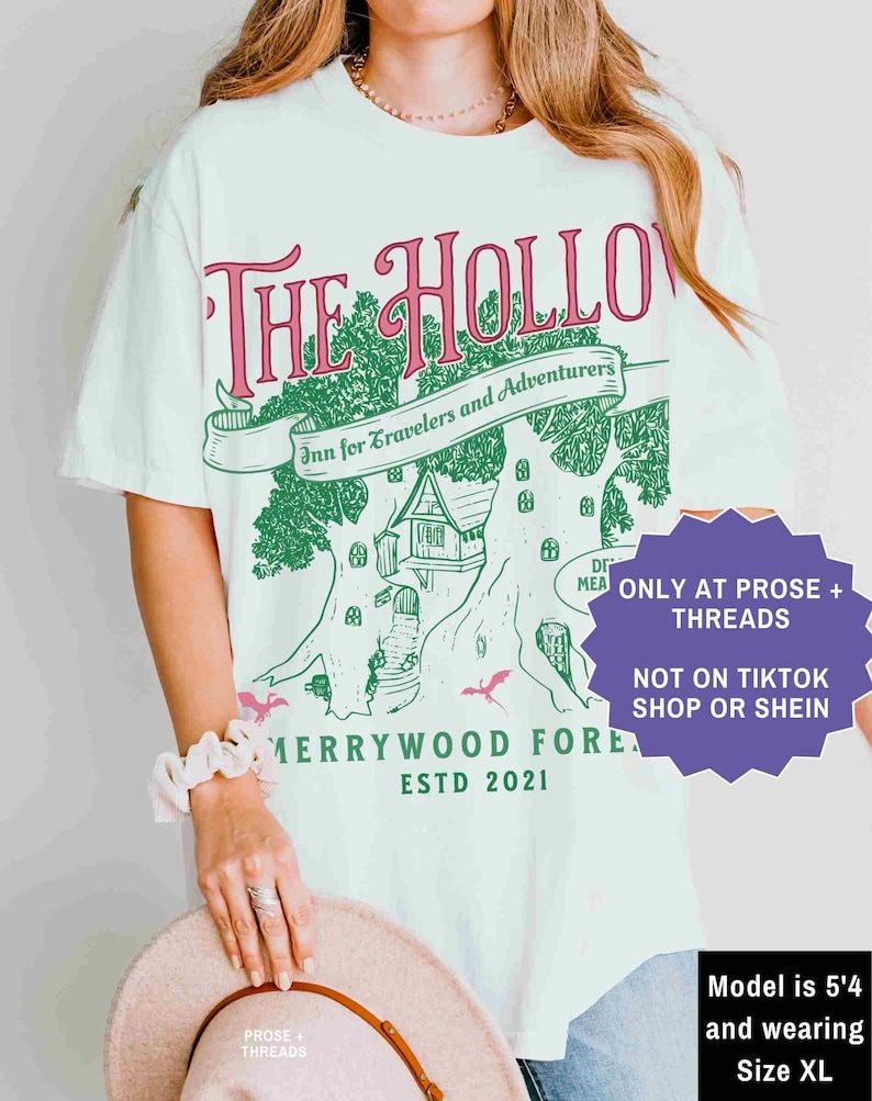 Pu&ograve; includere: T-shirt bianco con grafica verde e rosa con la scritta "The Hollow Inn for Travelers and Adventurers" con un disegno di casa sull'albero. La maglietta ha anche la scritta "Merrywood Forest ESTD 2021". La modella indossa la taglia XL.