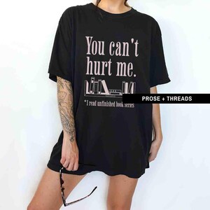 May include: Black oversized t-shirt with the text "You can't hurt me." and a graphic of a bookshelf with books. The text below the graphic reads "*I read unfinished book series". The text "PROSE + THREADS" is in the bottom right corner.