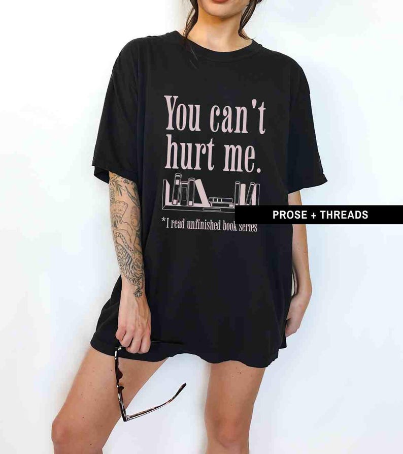 May include: Black oversized t-shirt with the text "You can't hurt me." and a graphic of a bookshelf with books. The text below the graphic reads "*I read unfinished book series". The text "PROSE + THREADS" is in the bottom right corner.