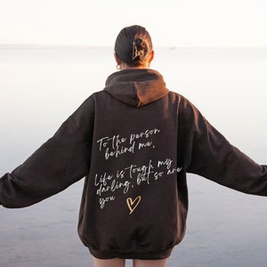 May include: A dark brown hoodie with the text "To the person behind me, Life is tough my darling, but so are you" in white script, along with a gold heart. The hoodie has a hood and long sleeves.
