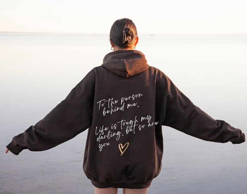 May include: A dark brown hoodie with the text "To the person behind me, Life is tough my darling, but so are you" in white script, along with a gold heart. The hoodie has a hood and long sleeves.