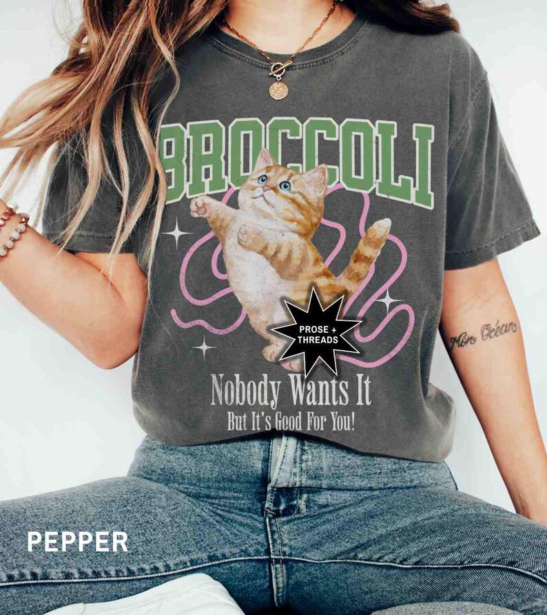 May include: A gray t-shirt with a green and white "BROCCOLI" graphic featuring a cartoon orange cat. The text "Nobody Wants It But It's Good For You!" is printed below the graphic. The t-shirt is from the brand "PROSE + THREADS".