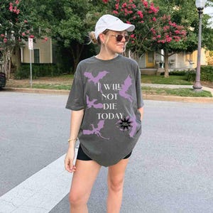 May include: A dark gray oversized t-shirt with a purple graphic of dragons and the text "I will not die today".