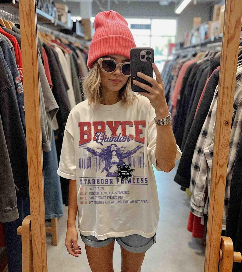 May include: A white t-shirt featuring a graphic print of a woman with the text "Bryce Quinlan" and "Starborn Princess". The shirt also includes lyrics from a song with the dates and titles of each line.