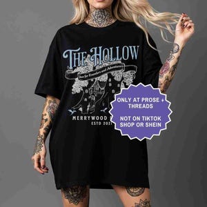 Pu&ograve; includere: T-shirt nero con un design grafico blu chiaro e bianco. Il design include il testo "The Hollow" sopra uno striscione che recita "Inn for Travelers and Adventurers". Sotto, un albero con una casa e il testo "Merrywood Estd 2021".
