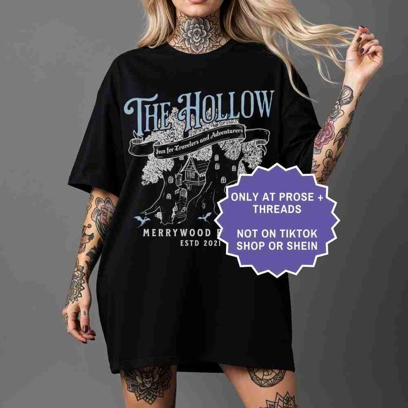 Pu&ograve; includere: T-shirt nero con un design grafico blu chiaro e bianco. Il design include il testo "The Hollow" sopra uno striscione che recita "Inn for Travelers and Adventurers". Sotto, un albero con una casa e il testo "Merrywood Estd 2021".