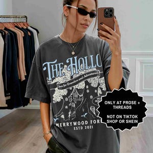Pu&ograve; includere: T-shirt grigio scuro con la scritta "THE HOLLO" in azzurro, con una grafica di casa sull'albero. La maglietta include anche il testo "Inn for Travelers and Adventurers" e "MERRYWOOD FOR ESTD 2021".