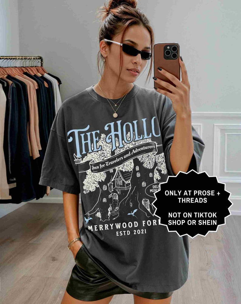 Pu&ograve; includere: T-shirt grigio scuro con la scritta "THE HOLLO" in azzurro, con una grafica di casa sull'albero. La maglietta include anche il testo "Inn for Travelers and Adventurers" e "MERRYWOOD FOR ESTD 2021".