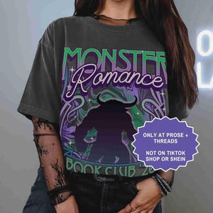 Op de afbeelding: Donkergrijs t-shirt met de woorden "Monster Romance Book Club '20" in groen en paars. De afbeelding toont een monstersilhouet. Een paars en wit label leest "ONLY AT PROSE + THREADS NOT ON TIKTOK SHOP OR SHEIN."