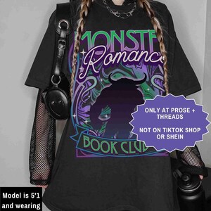 Op de afbeelding: Zwart oversized T-shirt met een afbeelding met de tekst "MONSTER Romance BOOK CLUB". Het ontwerp toont een silhouet van een monster met hoorns, en de tekst is in groen, paars en wit. Het shirt heeft korte mouwen.