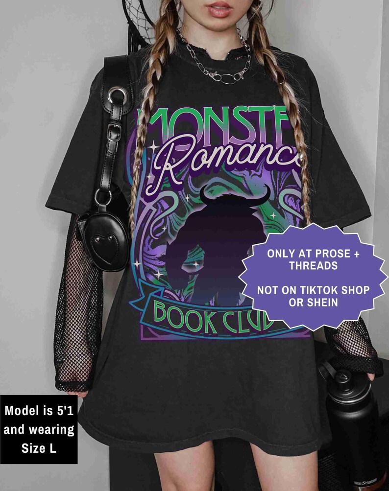 Op de afbeelding: Zwart oversized T-shirt met een afbeelding met de tekst "MONSTER Romance BOOK CLUB". Het ontwerp toont een silhouet van een monster met hoorns, en de tekst is in groen, paars en wit. Het shirt heeft korte mouwen.