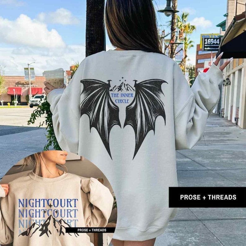 May include: Two cream colored sweatshirts with graphic designs. The first sweatshirt has a black bat wing design with the text "The Inner Circle" on the back. The second sweatshirt has a mountain range design with the text "Nightcourt Nightcourt Nightcourt" on the front.