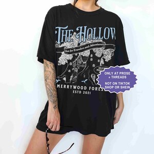 Pu&ograve; includere: T-shirt nera con una grafica con la scritta "THE HOLLOW" e "Merrywood Fores ESTD 2021". Il design include un albero con una casa e le parole "Inn for Travelers and Adventurers". La maglietta ha anche la scritta "ONLY AT PROSE + THREADS".