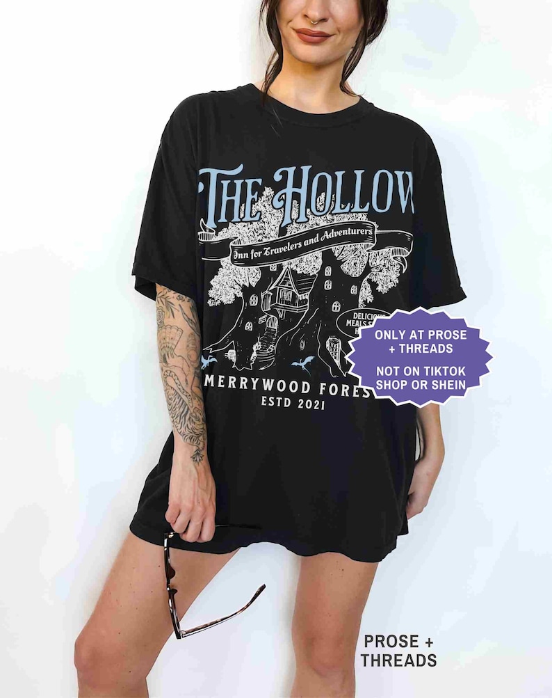 Pu&ograve; includere: T-shirt nera con una grafica con la scritta "THE HOLLOW" e "Merrywood Fores ESTD 2021". Il design include un albero con una casa e le parole "Inn for Travelers and Adventurers". La maglietta ha anche la scritta "ONLY AT PROSE + THREADS".