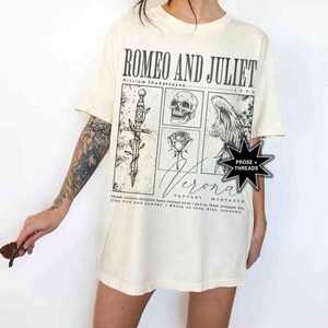 May include: Cream-colored t-shirt featuring a graphic design inspired by Shakespeare's "Romeo and Juliet." The design includes a sword, skull, rose, and a winged figure, with the text "Romeo and Juliet" and "Verona."